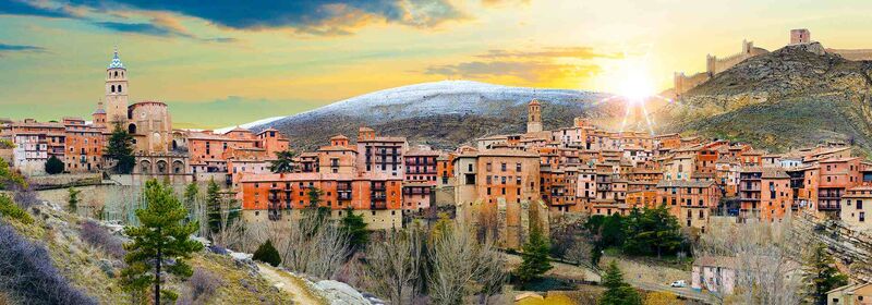 View of the medieval village Albarracin, in Spain. The historic city basks in the glow of the setting sun, with rooftops painted in warm hues and a backdrop of gentle hills.