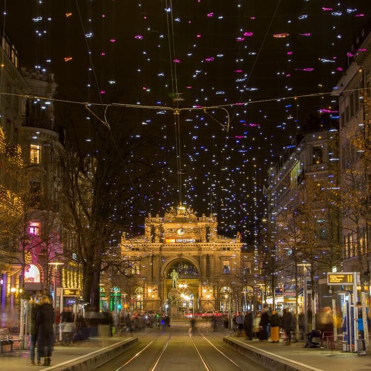 Nighttime scene in Zurich with colorful twinkling lights suspended above a bustling street, leading towards a grand illuminated building