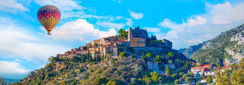 View of Eze, medieval town in France. The view shows the hilltop town crowned with ancient fortifications overlooks lush valleys, with a colorful hot air balloon floating in the bright sky.