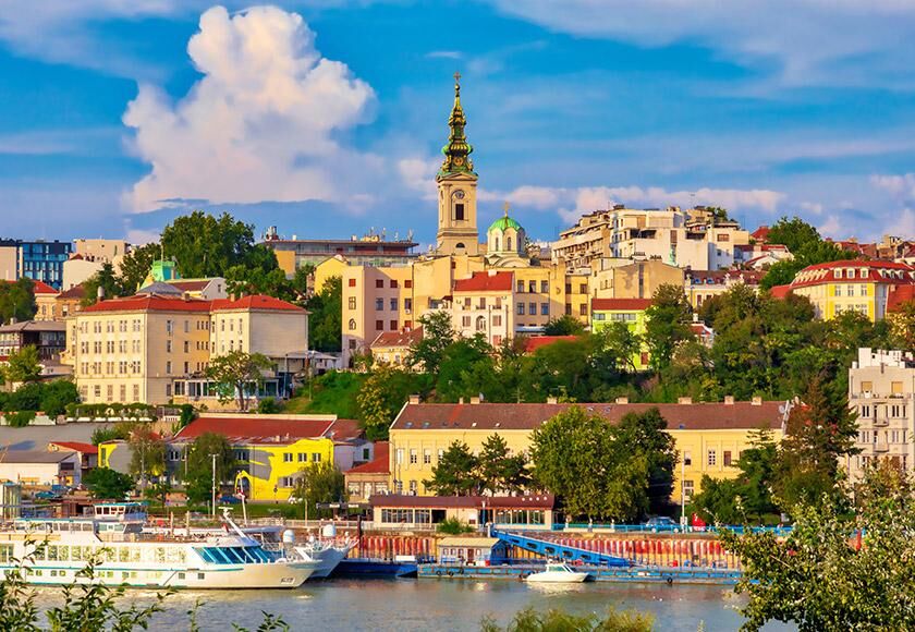 Belgrade city skyline with historic buildings, a prominent church tower, and boats anchored in the harbor under a clear blue sky