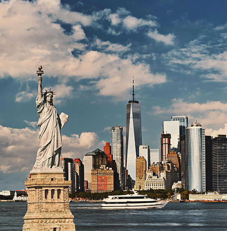 View of the Statue of Liberty with the New York skyline in the background against a blue sky with a few clouds.