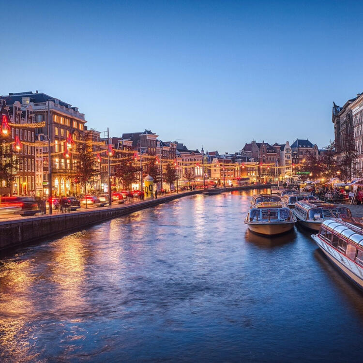 A serene evening view of Amsterdam canal, illuminated by city lights, with ornate buildings lining the waterside, tour boats docked, and a backdrop of a vibrant urban setting