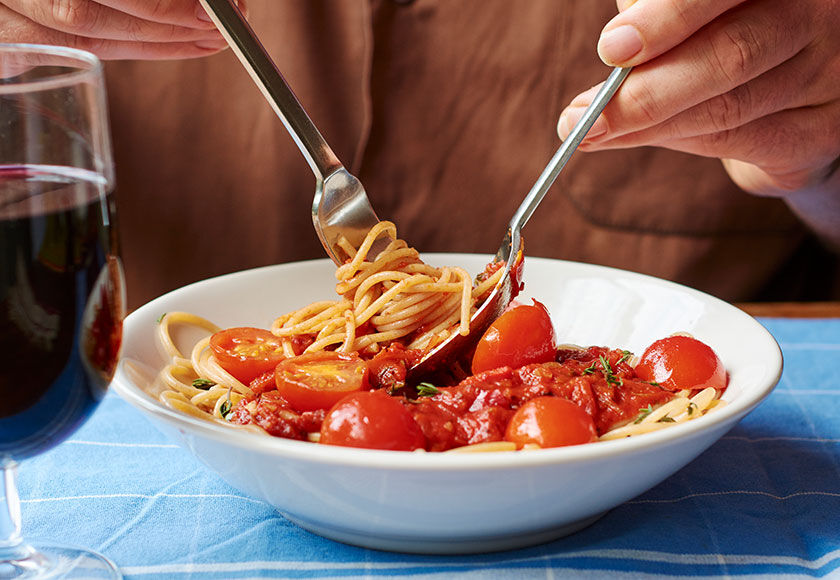 A person eating a dish of spaghetti with cherry tomatoes, cheese and spices, accompanied by a glass of wine