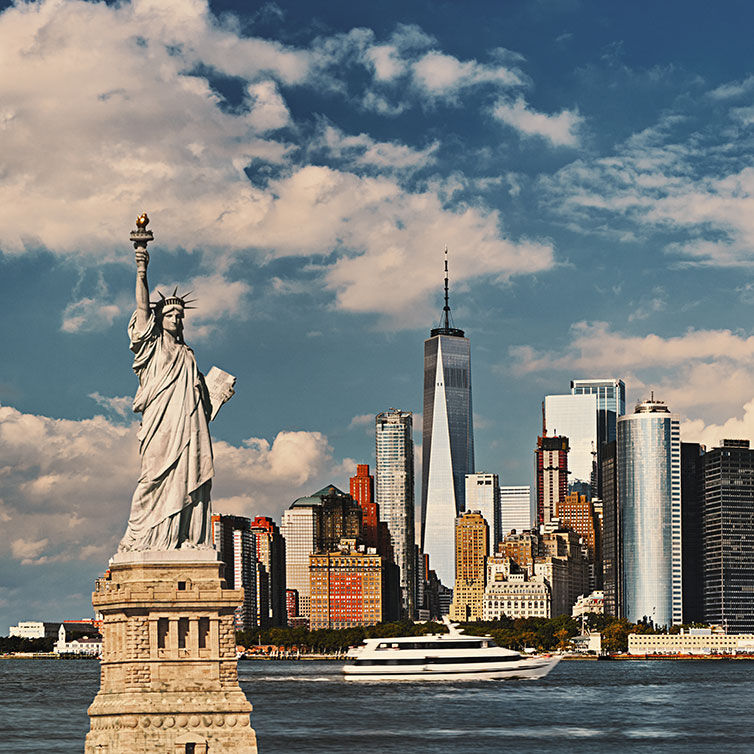 View of the Statue of Liberty with the New York skyline in the background, with a blue sky and a few clouds.