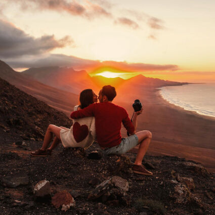A couple sitting on a hillside, embracing each other while watching a breathtaking sunset over a beach and the ocean, with the man holding a camera, capturing the moment.