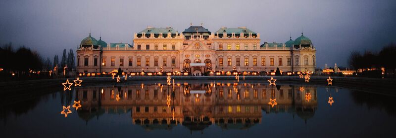 Overview of the Belvedere Palace in Vienna, with the Christmas Market, illuminated at twilight, reflecting symmetrically on calm waters against a dark blue sky.