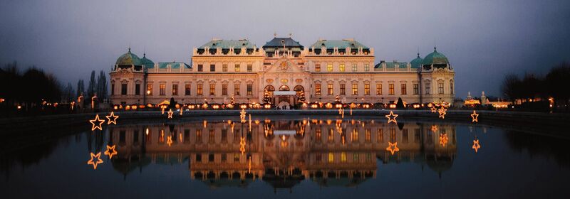 Flights to Vienna (VIE) Overview of the Belvedere Palace in Vienna, with the Christmas Market, illuminated at twilight, reflecting symmetrically on calm waters against a dark blue sky.