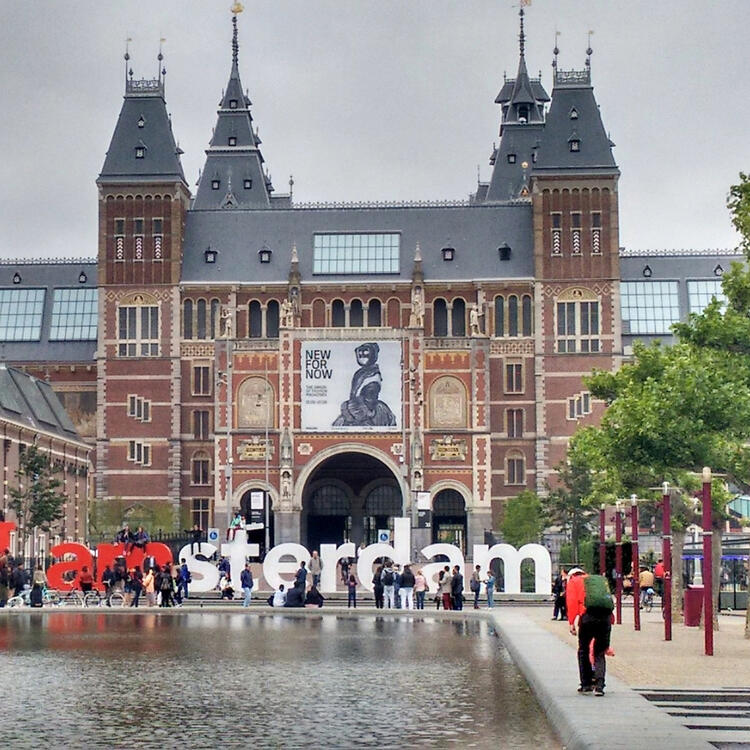 Iconic Amsterdam scene with the historic Rijksmuseum in the background, displaying an art banner, and the large 'I amsterdam' sign in the foreground, with tourists gathering around and a reflective water feature in front