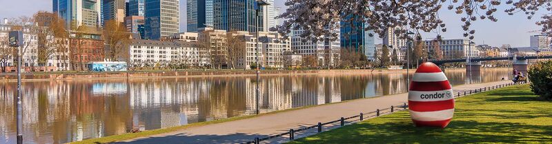 A red and white striped Easter egg with the Condor logo rests on the grass of a park with the city in the background.