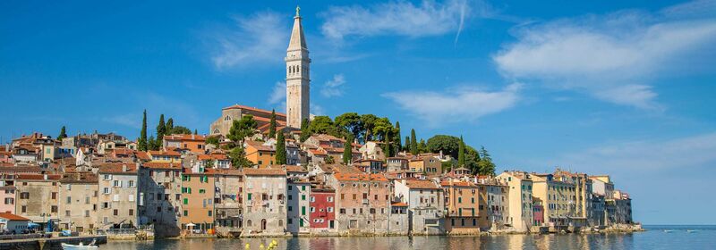 View of the Rovinj, in Croatia, a coastal town that graces the water's edge, with historic facades and a towering steeple set against a clear blue sky