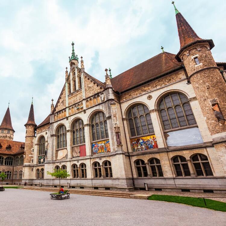 Stately gothic-style building in Zurich with intricate stone detailing, vibrant murals, pointed towers, and a person sitting on a bench in its spacious courtyard.