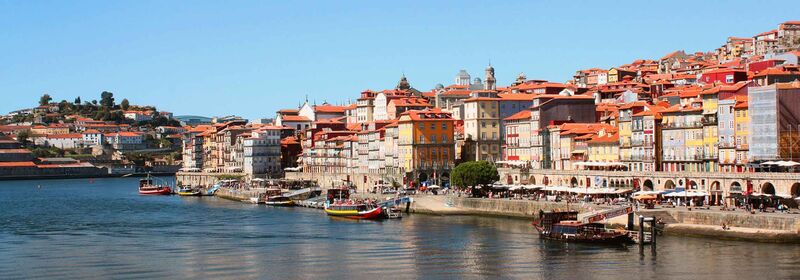 Flights to Porto (OPO) Porto and its picturesque riverside cityscape with colorful buildings, a calm river reflecting the town, and clear blue skies above.