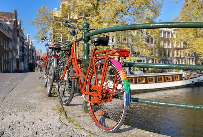 A red bicycle with the colors of the rainbow on the back is leaning against a canal railing in Amsterdam.