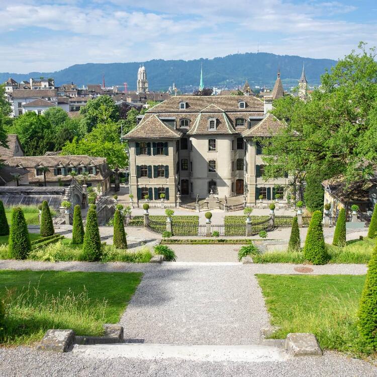 "Scenic view of a historic mansion with symmetrical gardens in Zurich, surrounded by lush greenery and with the city's skyline, including church spires and distant hills, in the background