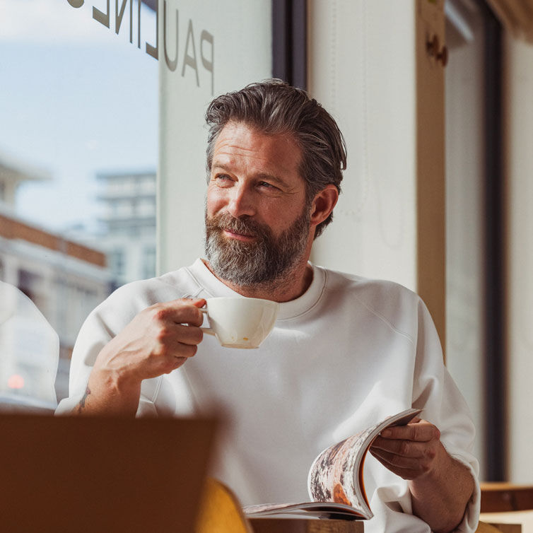 Man around 45 years old, with a beard and dark grey hair, drinking a coffee in an airport lounge