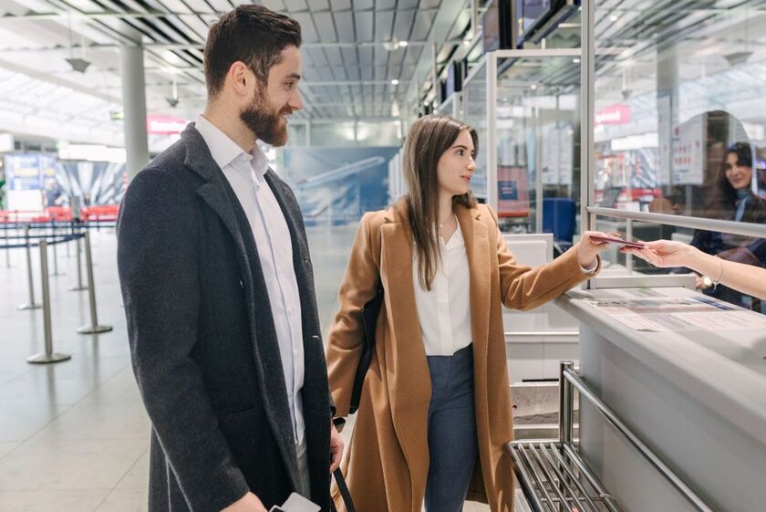 Man and woman in business-casual coats presenting travel documents at check-in counter in modern airport.