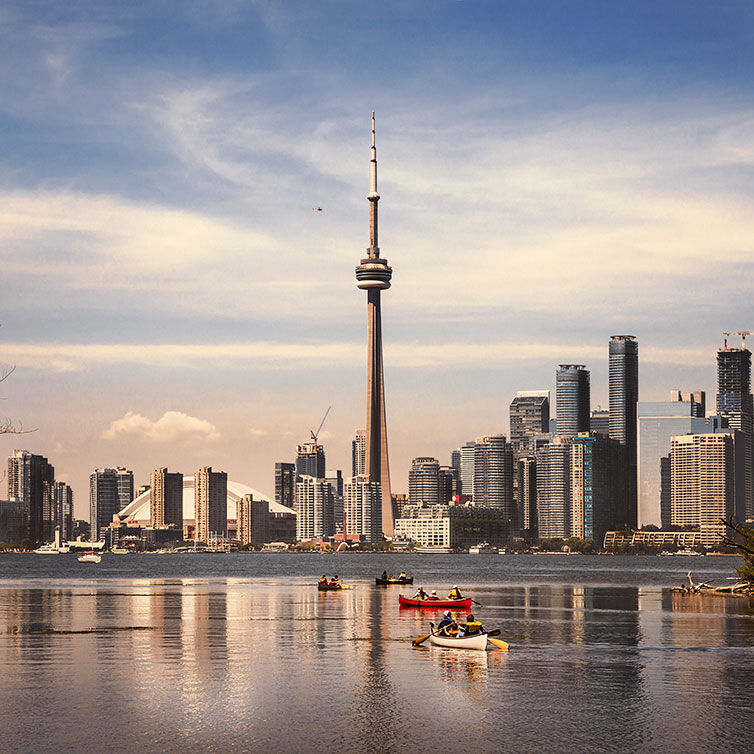 View of the Toronto skyline from the water, with the Canadian National Tower in the center. In the water are several boats with people paddling.