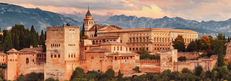 Alhambra fortress in Granada, Spain, bathed in warm sunlight with backdrop of cloudy blue skies and distant mountains.