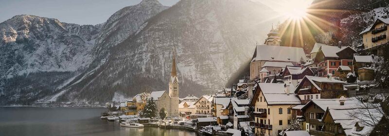 Hallstatt, in Austria, a historic village nestles by a lake, with alpine peaks rising behind it as the sun casts a golden glow over the snow-dusted landscape.