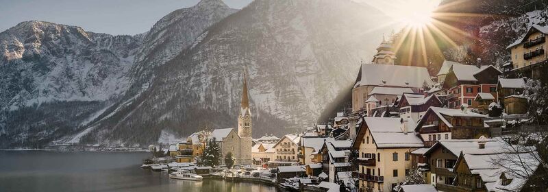 Hallstatt, Austria Hallstatt, in Austria, a historic village nestles by a lake, with alpine peaks rising behind it as the sun casts a golden glow over the snow-dusted landscape.